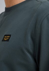 Tamnozeleni dukserica od mekane tkanine, sa okruglim izrezom, vidljivim šavovima i pravougaonom crnom etiketom sa zlatnim tekstom i logom.