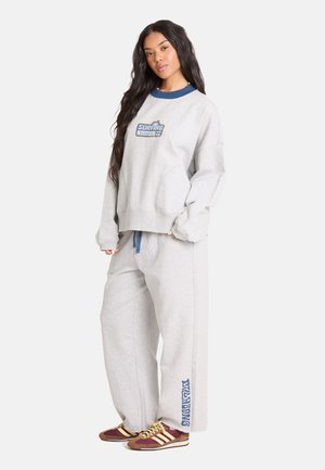 Jonge vrouw met lang zwart haar, gekleed in een lichtgrijze trui en wijde broek met surftekst en bruine sneakers.