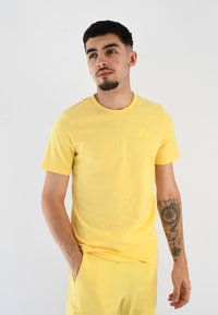 Camiseta de algodón de manga corta y cuello redondo en amarillo claro. Presenta un logotipo sutil en el pecho y una textura suave. Combinada con pantalones cortos a juego.