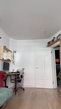 Ufficio domestico con una scrivania in legno dotata di computer, sedia in metallo e mensole a muro. Le pareti bianche e il pavimento in legno chiaro creano uno spazio aperto.