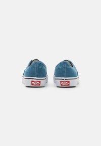 Chaussures slip-on en velours côtelé bleu avec une surface texturée, semelle en caoutchouc blanche et un accent de logo rouge au talon. Détails de couture circulaires visibles.