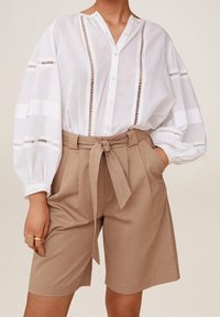 Valkoinen napitettava paita, jossa pystysuoria leikkauksia ja pussihihat, yhdistetty korkeavyötäröisiin beigeihin shortseihin, joissa on solmittava vyö.