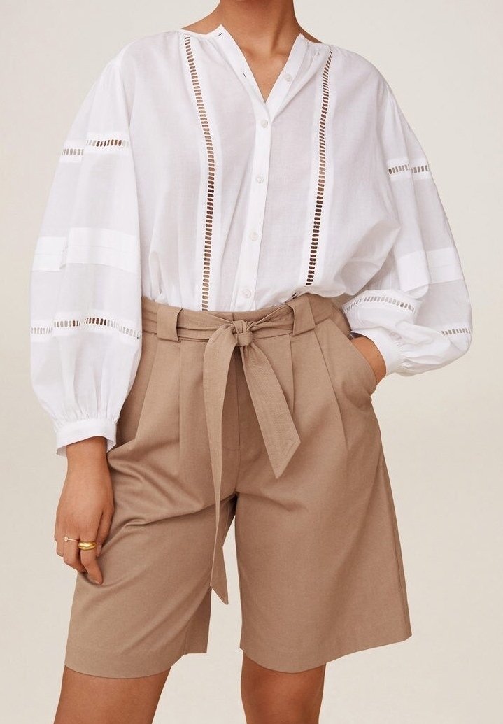 Valkoinen napitettava paita, jossa pystysuoria leikkauksia ja pussihihat, yhdistetty korkeavyötäröisiin beigeihin shortseihin, joissa on solmittava vyö.