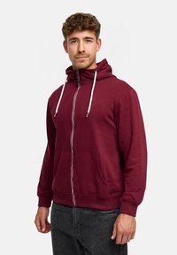 Burgundy zip-up hoodie made of cotton blend, featuring a drawstring hood, kangaroo pockets, white zippers, and ribbed cuffs.