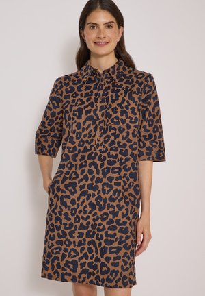 Kvinde iført en brun og sort leopardprint kjole med trekvartlange ærmer, krave og knapper foran, står mod en ensfarvet baggrund.