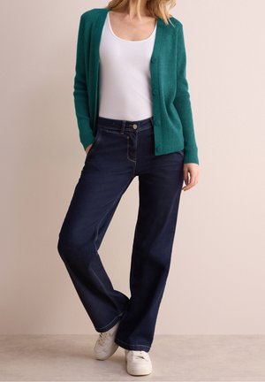 Personne portant un cardigan turquoise boutonné, un top blanc à encolure dégagée, un jean large bleu foncé, et des baskets blanches, debout les mains dans les poches.
