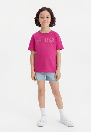 Child standing in pink GUESS T-shirt, light blue denim shorts, white socks, and white sneakers, against a plain white background.