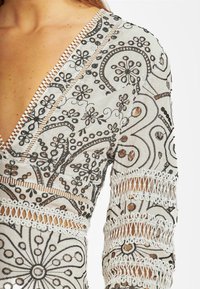 Vestido bordado con un escote en V profundo, que presenta intrincados patrones negros sobre una tela clara, y un delicado borde de encaje blanco en las mangas.