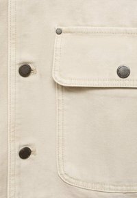 Beige canvasjas met een klepzak, zilverkleurige metalen knopen en stikdetails. De stof heeft een stevige, gestructureerde uitstraling.