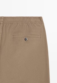 Brązowe spodnie z tkaniny o splocie, z elastycznym pasem i tylną kieszenią zapinaną na guzik, ukazujące gładką fakturę i dopasowany krój.