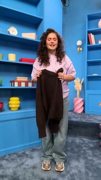 Frau mit lockigem Haar steht in einem blauen Zimmer und hält einen dunkelbraunen Pullover hoch, umgeben von bunten Regalen mit Büchern und Dekoration.