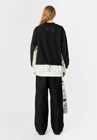 Sudadera de gran tamaño en negro y crema con dobladillo texturizado, que cuenta con un bolsillo trasero con solapa, combinada con pantalones amplios negros.