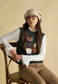 Zaļš adīts džemperis ar izšūtu lapsas dizainu, valkāts virs baltas krekla ar apkakli un mežģīņu apdari, kopā ar raibu brūnu biksēm.