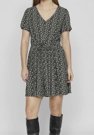 Woman wearing a short black dress with white leaf pattern, v-neck, short sleeves, and black knee-high boots against plain background.