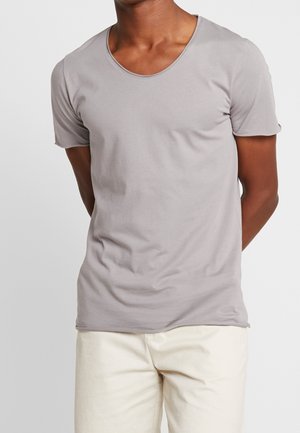 Mężczyzna w jednokolorowej, jasnoszarej koszulce z krótkim rękawem i kremowych spodniach, stojący z rękami za plecami na jednolitym tle.