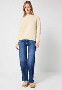 Maglione beige a coste con maniche lunghe, abbinato a jeans blu a gamba dritta e stivaletti beige, su uno sfondo neutro.