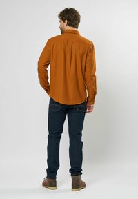 Gebrand oranje lange mouwen shirt met een spitse kraag, achterkant plooidetail en knoopmanchetten, gecombineerd met donkerblauwe jeans en bruine laarzen.
