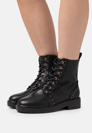 Bottes cheville en cuir noir avec œillets dorés, fermeture à lacets sur le devant et accents cloutés sur les côtés. Semelle plate et bout arrondi.