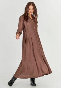 Long brown shirt dress made of a textured fabric with a buttoned front, collar, and long sleeves, paired with black ankle boots.