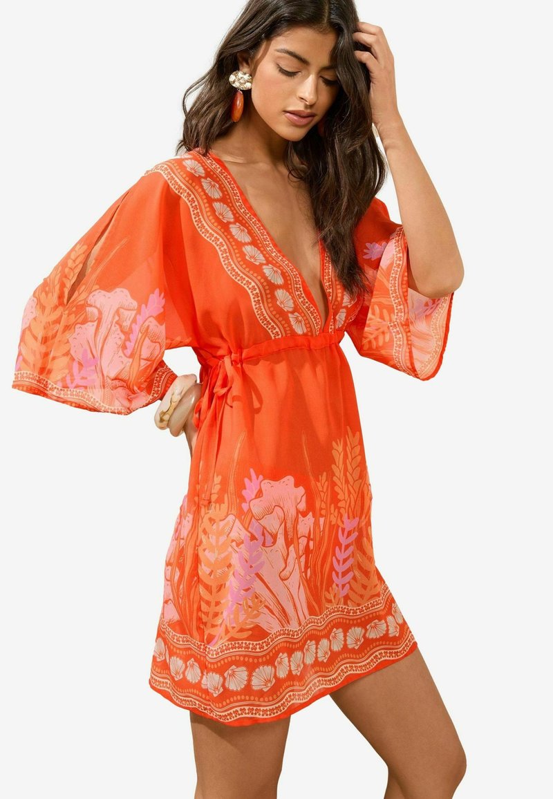 Svetlooranžový kaftan s véčkovým výstrihom a širokými rukávmi, s kvetinovými motívmi v ružovej farbe. Obsahuje pás na zaväzovanie a dekoratívny lem na spodnom okraji.