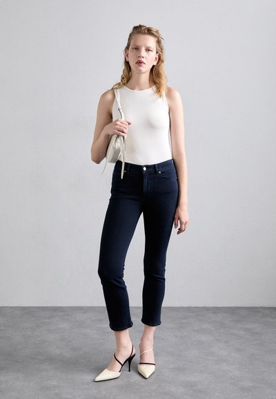 Bodysuit sans manches blanc, jeans skinny bleu marine avec une légère élasticité, et escarpins beiges à bout pointu avec des brides noires autour de la cheville. Sac à main minimaliste.