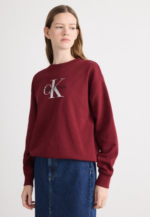 Burgundy sweatshirt with a crew neckline, featuring a large silver "CK" logo. Worn with a denim skirt, showcasing long sleeves and a relaxed fit.