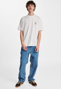 Biała koszulka bawełniana z małą zieloną grafiką po lewej stronie klatki piersiowej, w połączeniu z luźnymi niebieskimi dżinsami i brązowo-czarnymi trampkami.