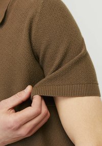 Barna, textúrás pólóing rövid ujjakkal. A közeli felvételen a bordázott mandzsetta látható, és egy kéz a ujj rögzítésénél, kiemelve az anyag részleteit.