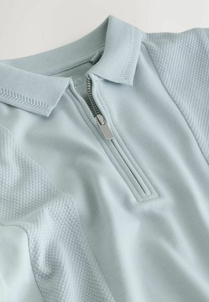 Svetlo modra pulover z zadrgo na ovratniku, z teksturiranimi pletenimi paneli na ramenih in gladkim tkanim materialom na telesu.