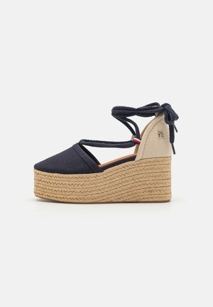 Espadrille compensée en toile marine avec des brides croisées autour de la cheville, une semelle en jute tressé, et un panneau accent beige arborant un petit logo.