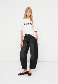 Λευκό γραφικό t-shirt με τη λέξη "LOVE" σε μαύρο και κόκκινο, συνδυασμένο με ψηλόμεσα σκούρα τζιν. Το μοντέλο φοράει γυαλιστερά μπορντό πλατφόρμες.
