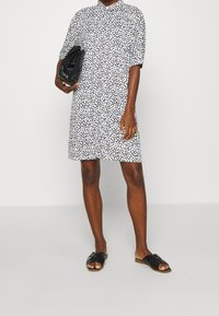 Robe chemise à manches courtes blanche avec un motif à pois noirs, col classique et coupe ample. Associée à des sandales plates noires et à une pochette.