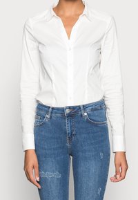 Kvinna iförd en åtsittande vit skjorta med knappar, instoppad i blå denimjeans med mindre slitage på benen.