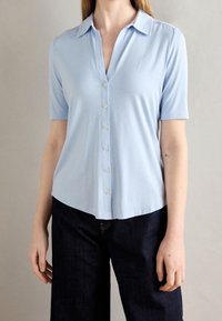 Camicia azzurro chiaro a maniche corte con colletto, chiusura frontale con bottoni e orlo leggermente curvo. Tessuto liscio senza motivi.