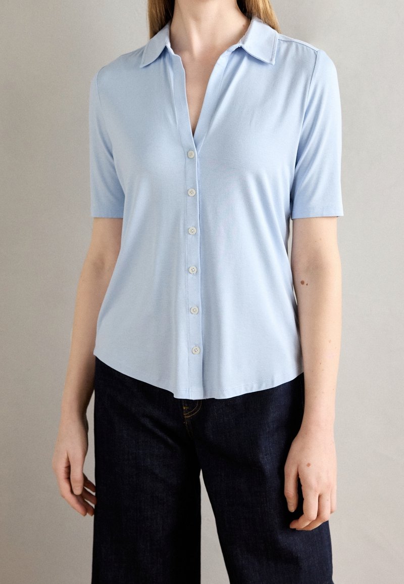 Camicia azzurro chiaro a maniche corte con colletto, chiusura frontale con bottoni e orlo leggermente curvo. Tessuto liscio senza motivi.
