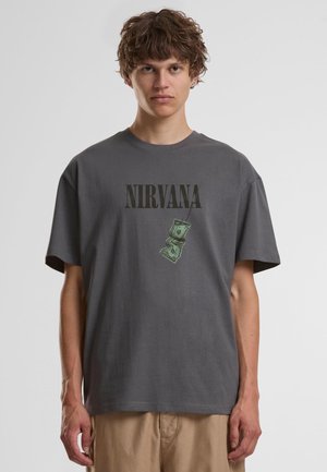 Jeune personne aux cheveux bouclés portant un t-shirt Nirvana gris foncé et un pantalon beige sur un fond clair uni.