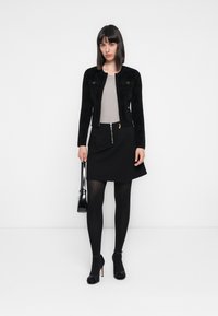 Donna che indossa una giacca nera, un top beige, una gonna nera con cerniera, collant neri, tacchi neri e tiene una borsa nera su uno sfondo bianco.