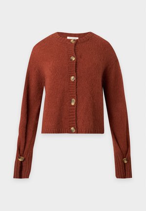 Femme debout avec une main dans la poche, portant un cardigan rouille boutonné, un jean noir à jambes larges et des talons ouverts noirs sur fond blanc.