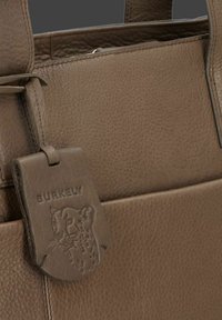 Bruine leren tote bag met een gestructureerde afwerking, voorzien van een leren label met een reliëf ontwerp van een luipaard en het merk "BURKELY."