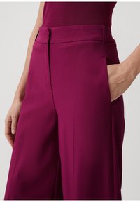 Pantalons évasés en tissu magenta riche, dotés de poches latérales, d'une taille plate et d'une texture lisse.