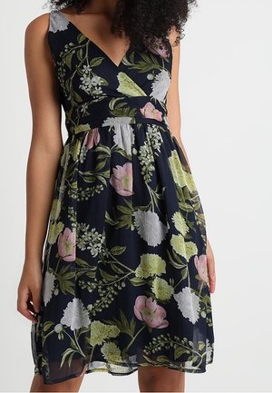 Robe noire sans manches avec motif floral rose, blanc et vert, encolure en V, taille ajustée et jupe évasée portée par une femme.
