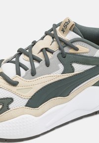 Lightweight athletic shoe with a mesh upper, beige and green accents, and textured overlays. Features rounded toe and thick white sole.