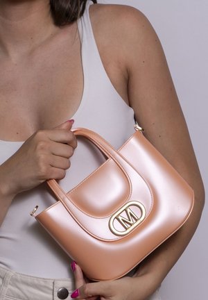 Femme tenant un petit sac à main pêche brillant avec un fermoir ovale doré portant la lettre "M", portant un haut blanc sans manches.