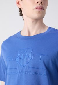 Mėlynas medvilnės marškinėliai su iškilusiu GANT logotipu ir tekstu "1949 AMERICAN SPORTSWEAR" kontrastinga mėlyna siūle.