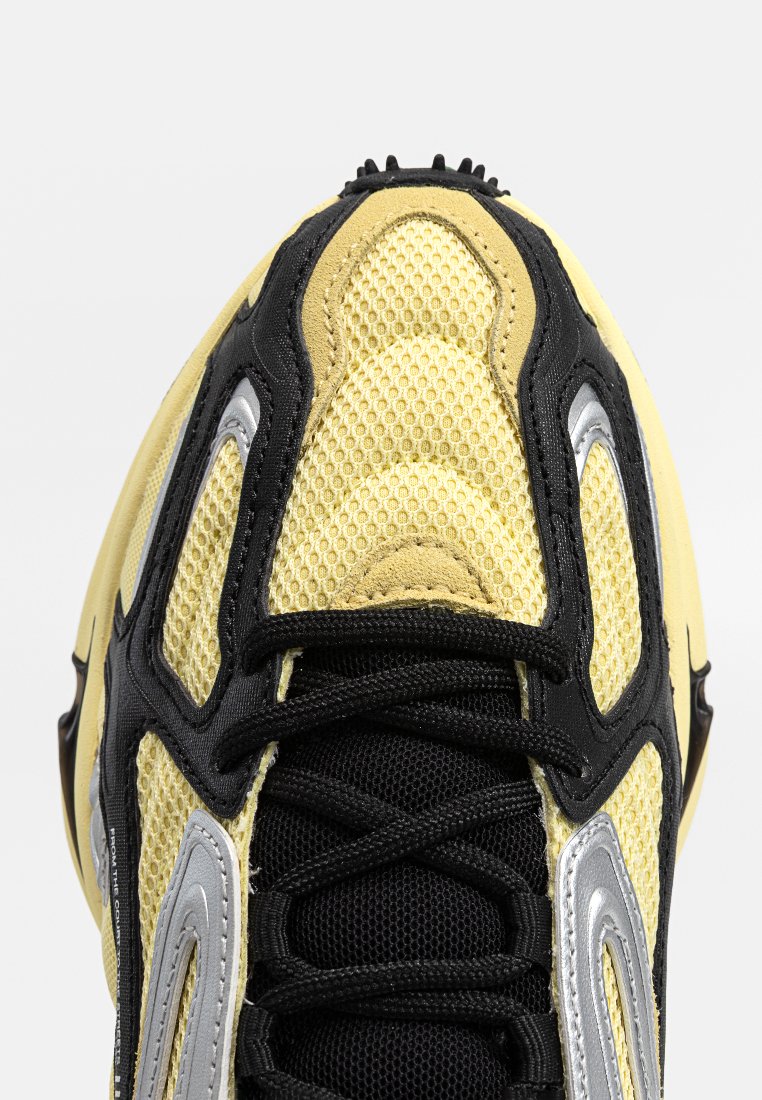 Buty sportowe z siateczkową cholewką w kolorze żółtym i czarnym, z dodatkami w kolorze srebrnym, teksturowanymi wzorami i czarnymi sznurówkami zapewniającymi pewne dopasowanie.