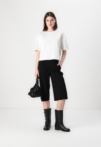Tricou alb scurt, pantaloni culotte negri cu croială lejeră, cizme negre până la genunchi cu nasturi și o geantă modelată neagră. Design minimalist.