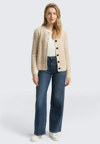 Beige, flauschiger Cardigan mit schwarzen Knöpfen, gerippten Bündchen und Saum, kombiniert mit hochtaillierten blauen Jeans und weißen Turnschuhen.