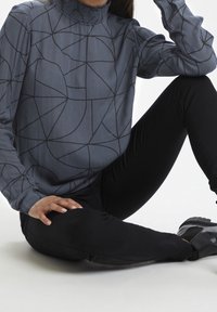 Langærmet blå bluse med sort geometrisk mønster og rynket krave, parret med sorte bukser og sko.