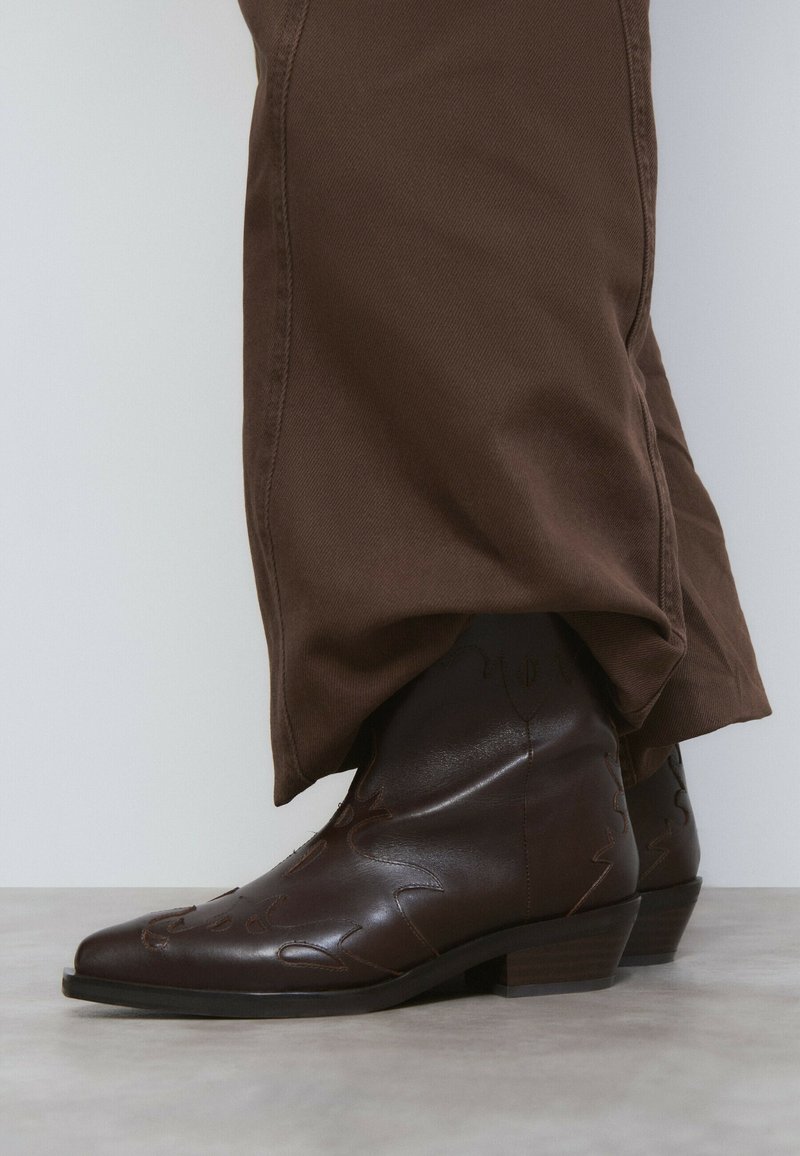 Botas de vaquero de cuero marrón con punteras puntiagudas y costuras decorativas, combinadas con pantalones marrones sueltos. Textura suave y tacones bajos en bloque.