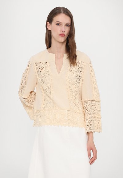 Beige bluse med blonderaccentpaneler, featuring en V-hals, trekvartærmer og en scalloped kant. Blødt stof med intrikate mønstre.
